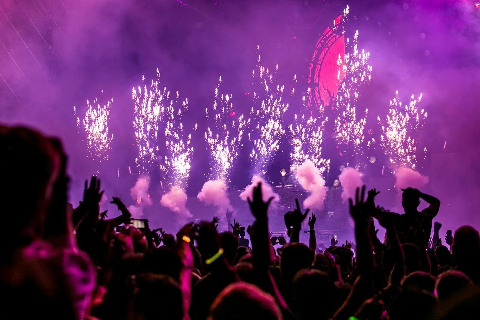 Excited crowd with raised hands cheering at a live music performance under colorful stage lights