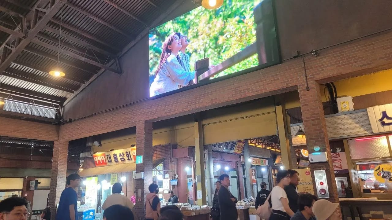 Large screen above the food court inside Yesan Traditional Market with shops on either side